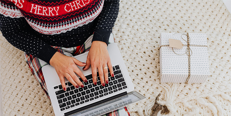 Overhead view of person wearing a Christmas jumper sitting with laptop on their lap. A present is sitting next to them.